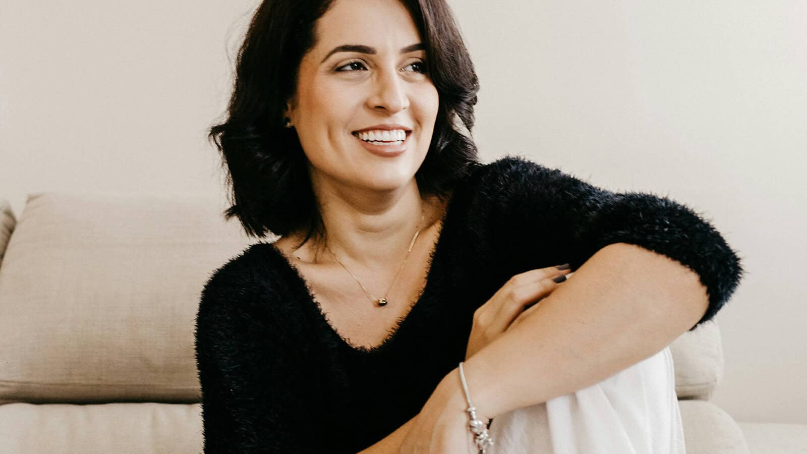 A woman with dark hair and a black sweater smiling while looking off-camera.