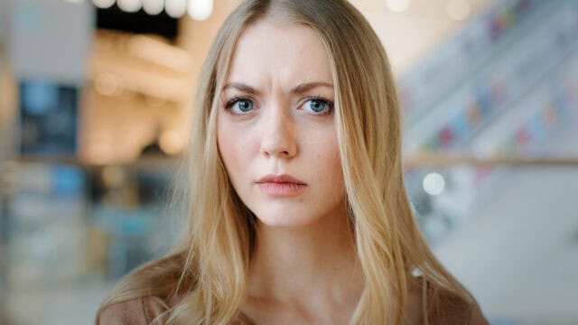 A young woman with long blonde hair stares directly at the camera with a serious, slightly concerned expression. The background is softly blurred, suggesting an indoor setting.