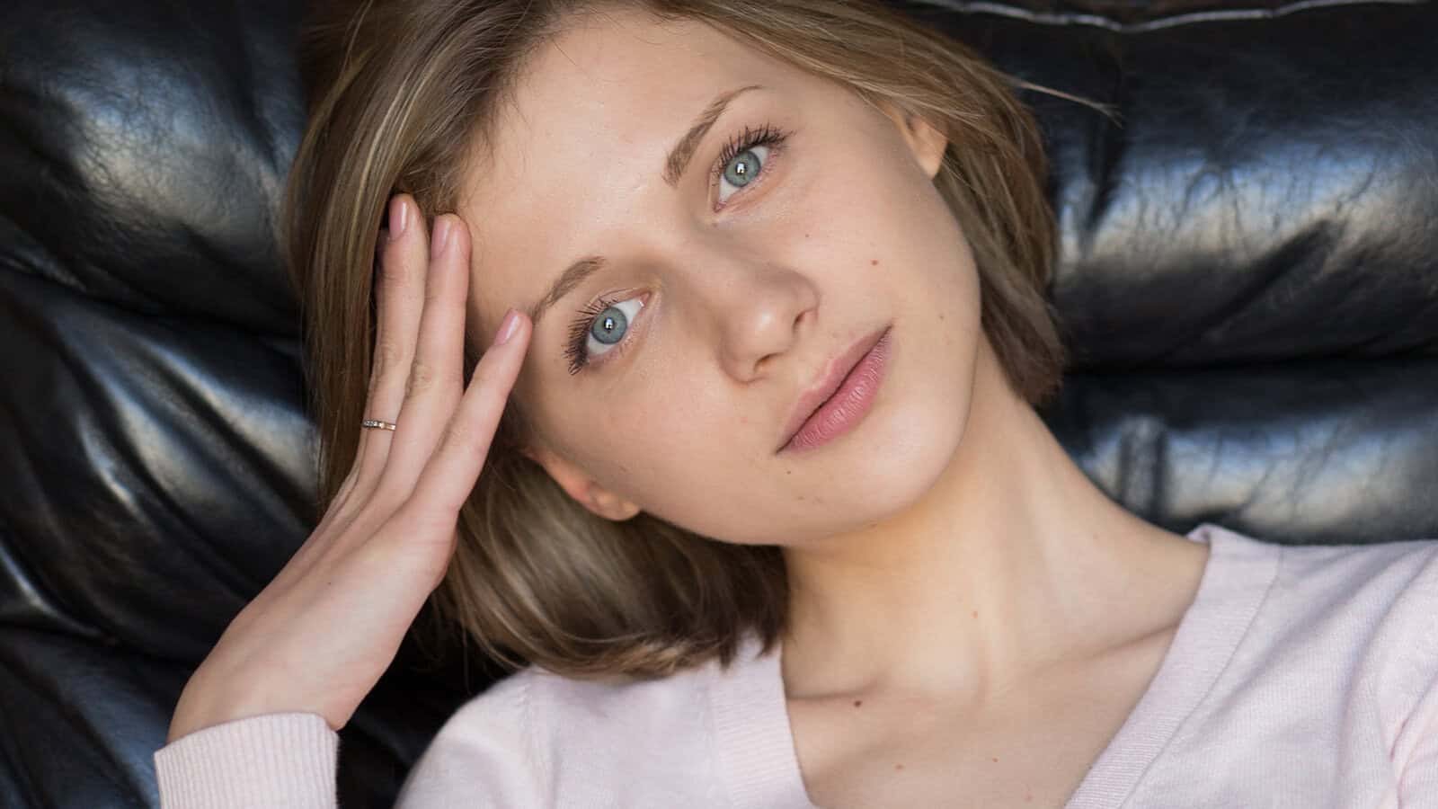 A young woman with light brown hair and blue eyes looking toward the camera with her hand resting against her forehead.