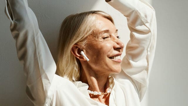 A smiling woman with blonde hair wearing wireless earbuds and reaching her arms upward.