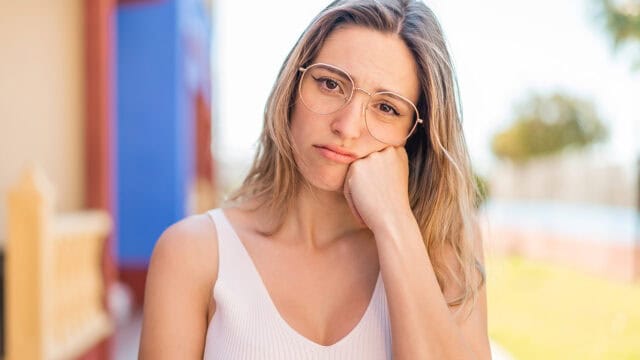 A woman wearing glasses rests her cheek on her hand with a pensive expression.