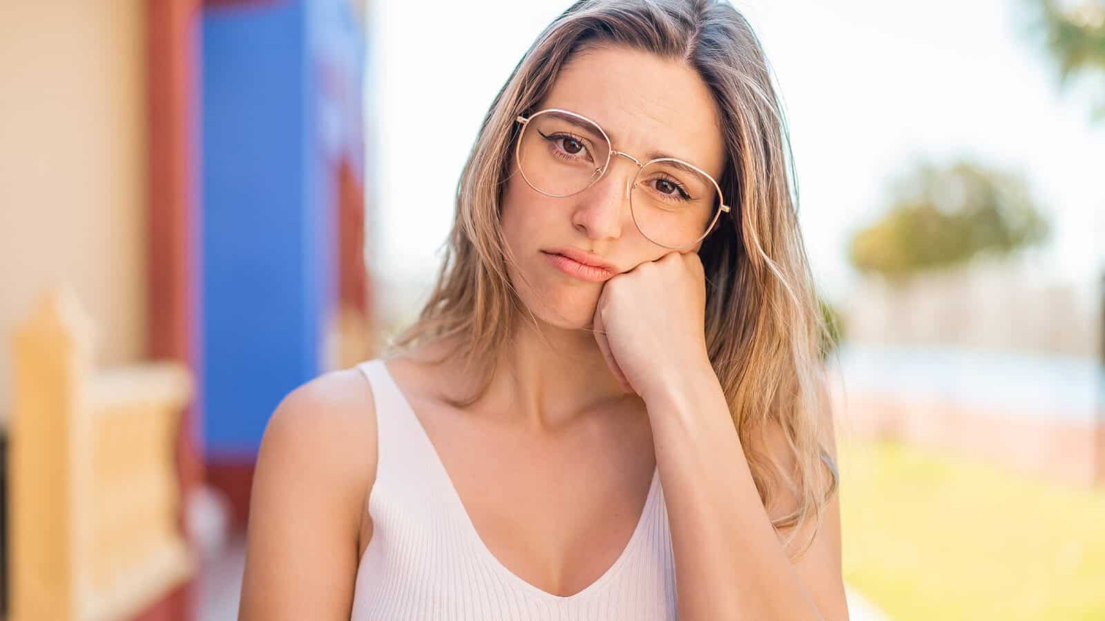 A woman wearing glasses rests her cheek on her hand with a pensive expression.