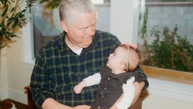 A man in a plaid shirt holds a baby, looking at each other affectionately.