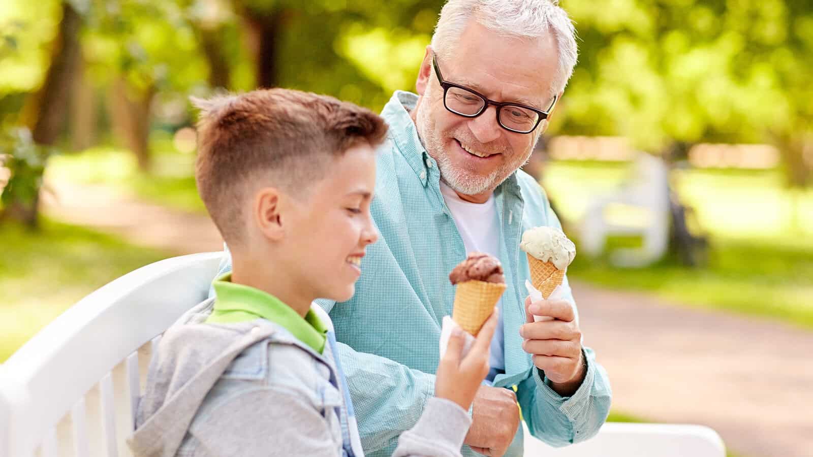An older man and a young boy sitting on a park bench, both smiling while holding ice cream cones.