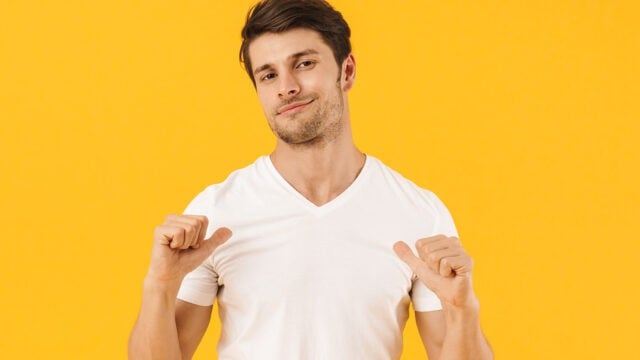 A man in a white t-shirt points both thumbs toward himself and smiles slightly, standing against a bright yellow background.