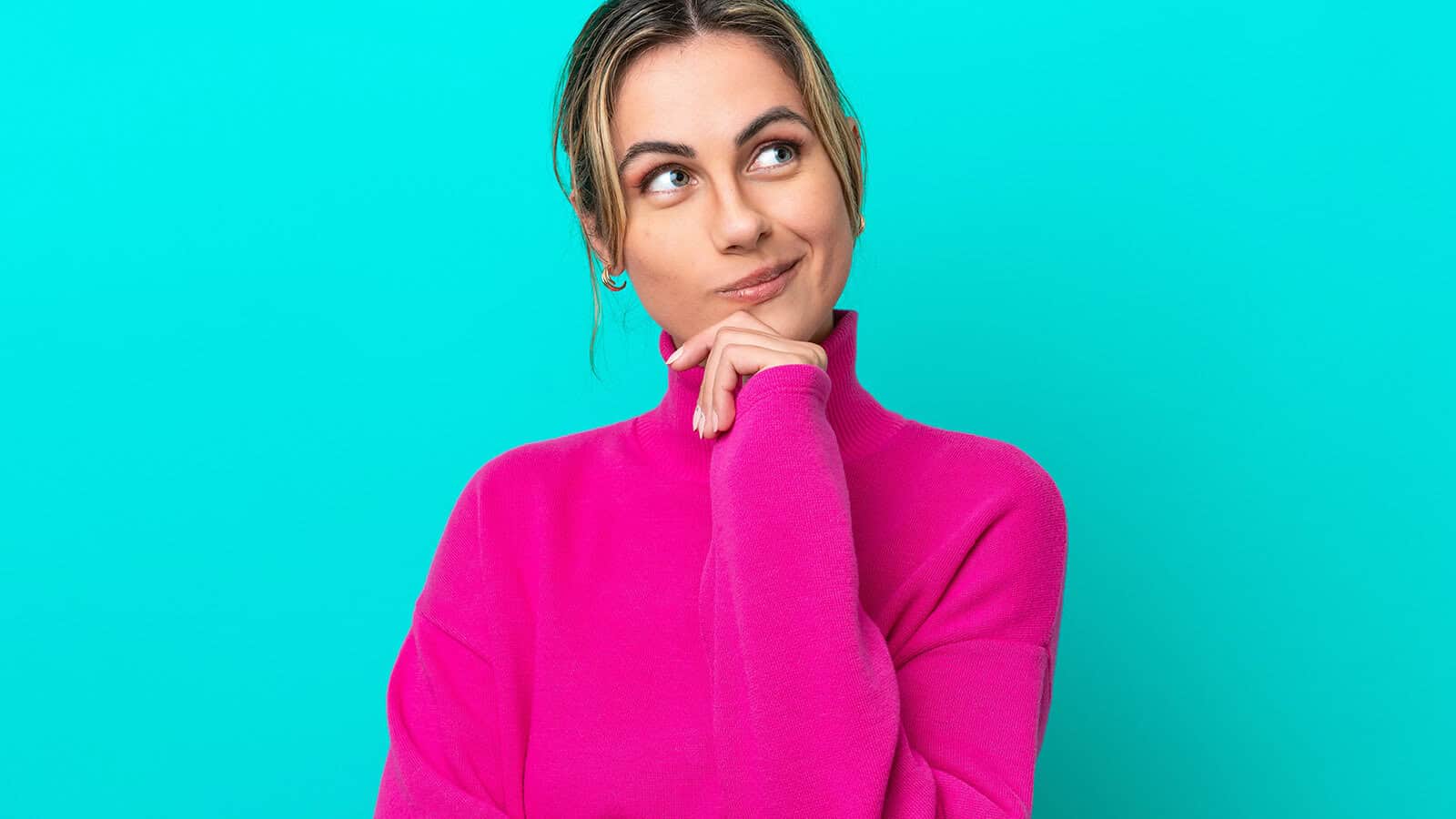 A person in a bright pink turtleneck sweater looking upward with a thoughtful expression, resting their chin on their hand.