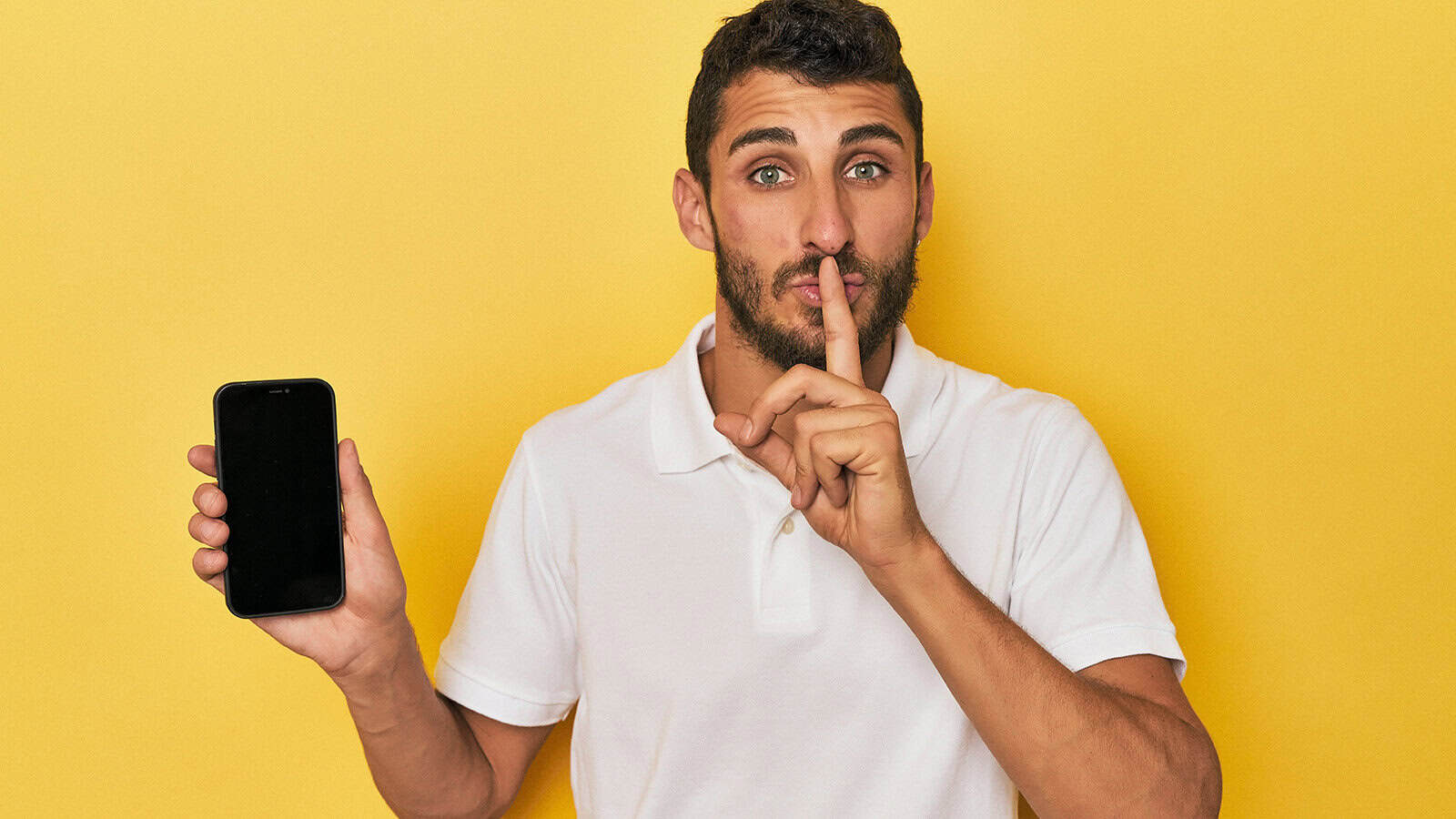 A man in a white polo shirt holds up a smartphone while making a shushing gesture with his finger against his lips.