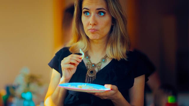 A woman holding a small paper plate of food looks to the side with an surprised or curious expression.