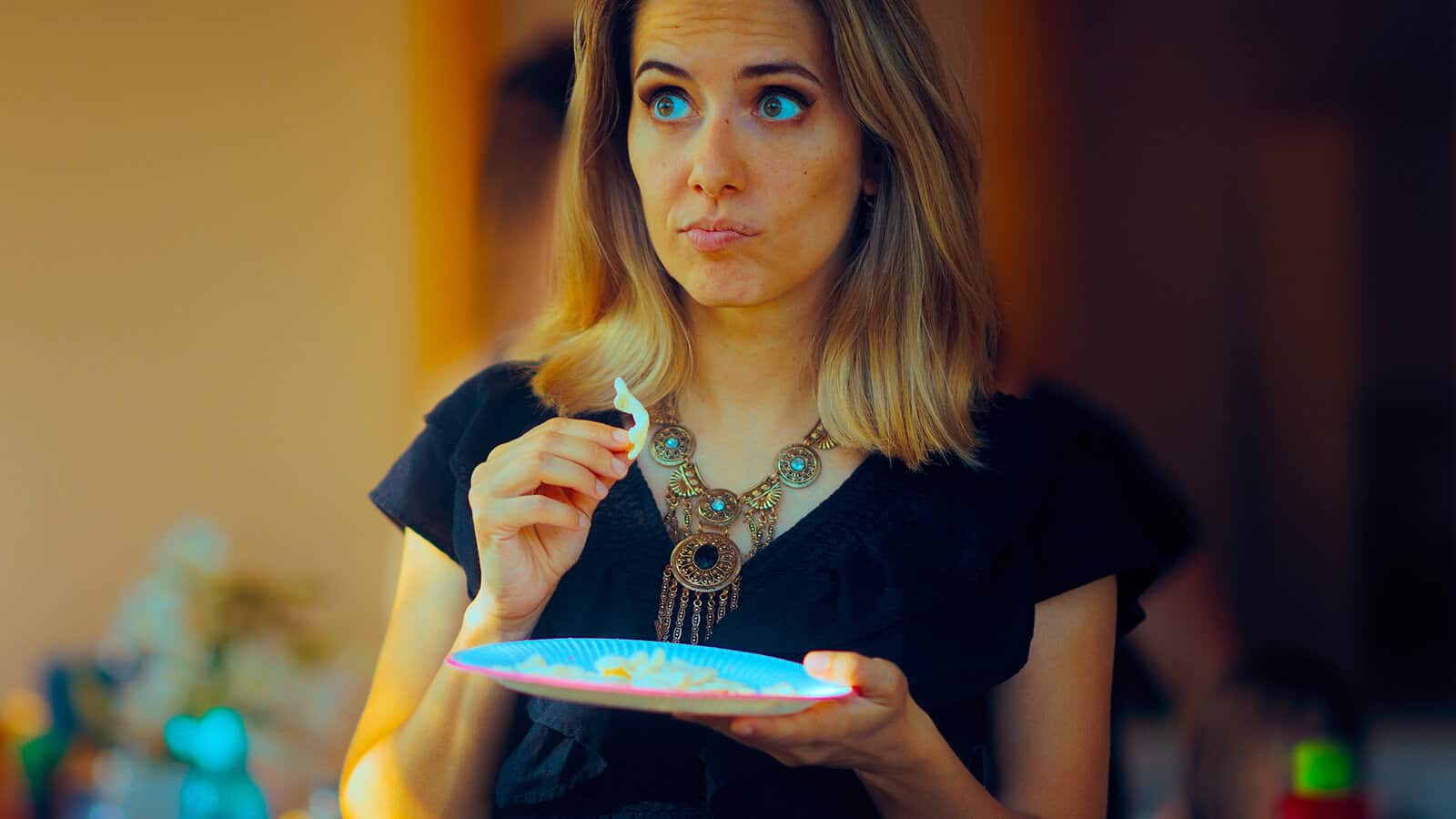 A woman holding a small paper plate of food looks to the side with an surprised or curious expression.