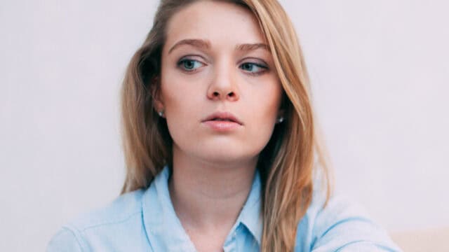 A young woman with long blonde hair, wearing a light blue shirt, looks to her left with a neutral expression against a plain light background, lost in self-reflection.