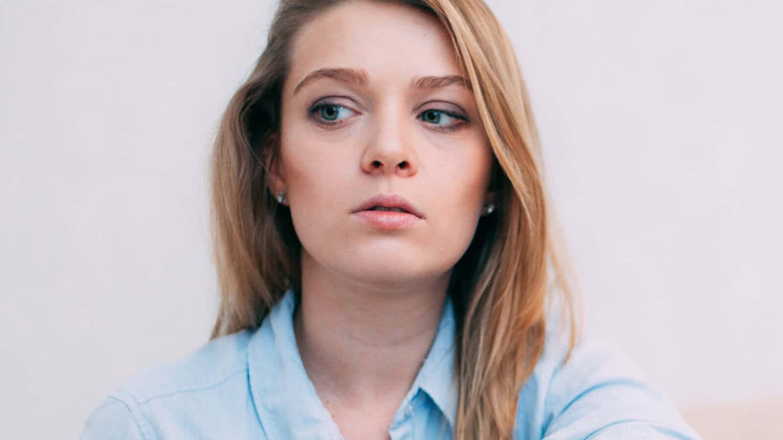 A young woman with long blonde hair, wearing a light blue shirt, looks to her left with a neutral expression against a plain light background, lost in self-reflection.