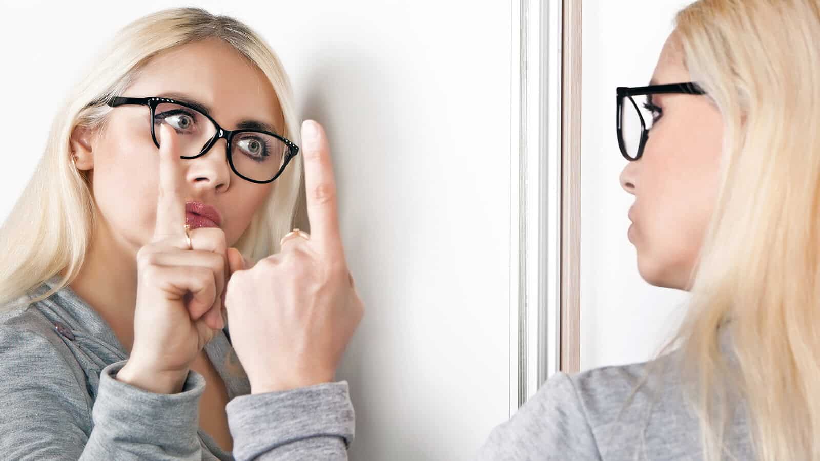 A blonde woman wearing glasses looks at herself in a mirror, holding up her index finger and pressing it against her lips in a shh gesture, suggesting silence or secrecy.