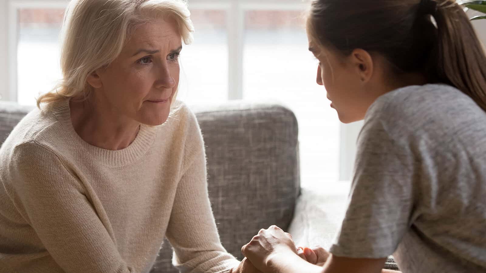 An older woman and a younger woman sit together, holding hands while having a serious conversation.