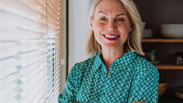 A smiling woman with blonde hair wearing a green geometric-print collared shirt standing next to window blinds.