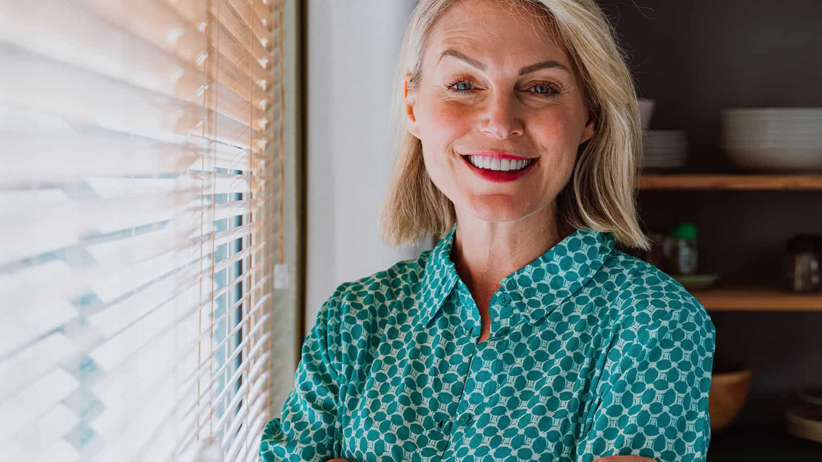 A smiling woman with blonde hair wearing a green geometric-print collared shirt standing next to window blinds.