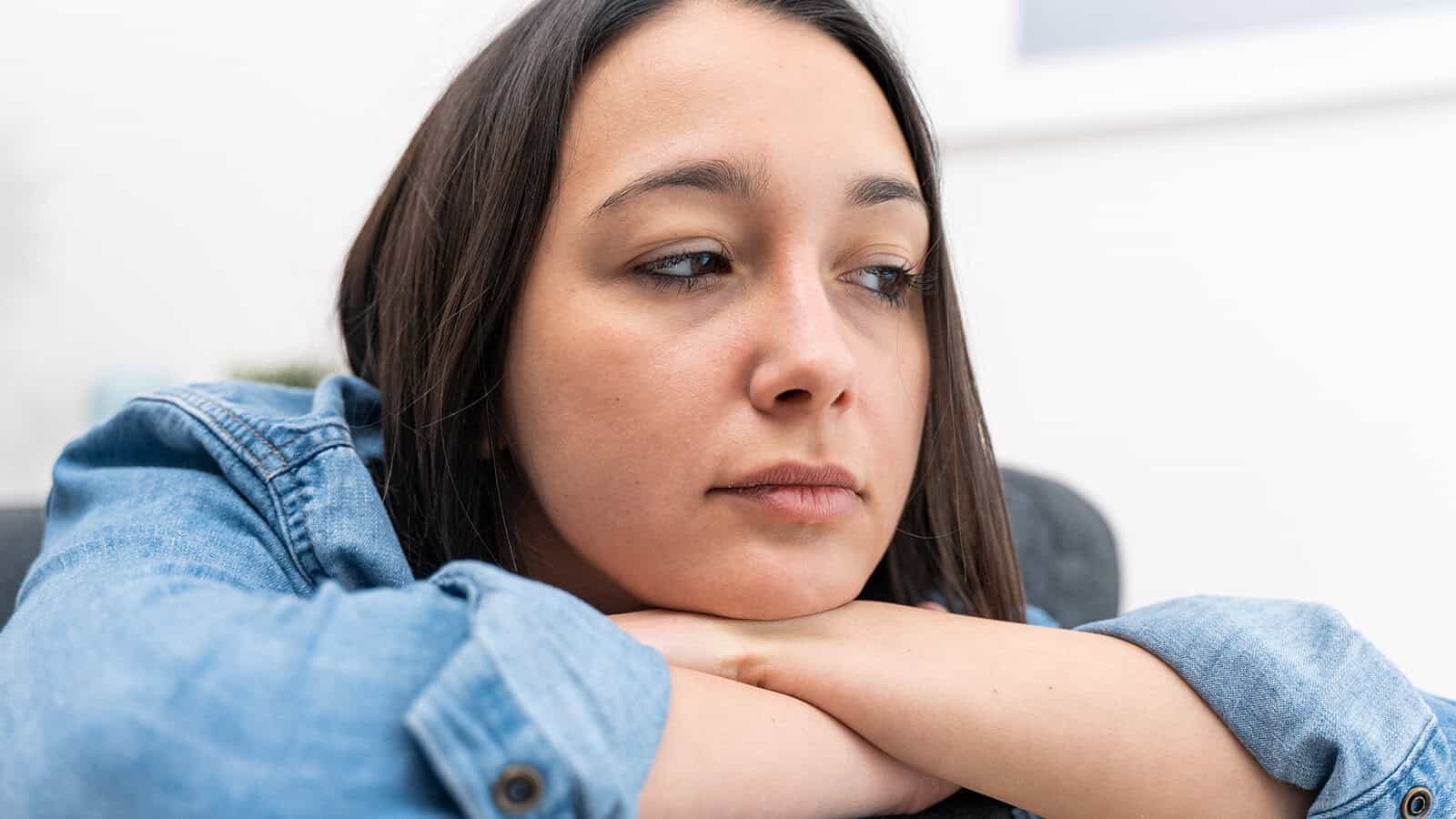 A young woman with dark hair leans her chin on her hands, looking away with a pensive expression.