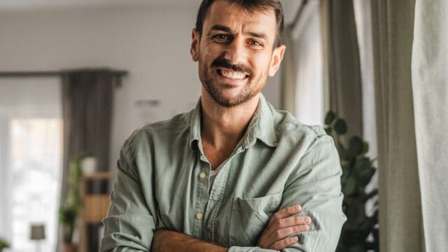 A smiling man with a beard wearing a light green button-down shirt stands with his arms crossed.