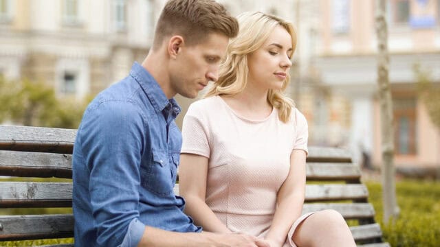 A man and a woman sitting side by side on a park bench, looking down with somber expressions.