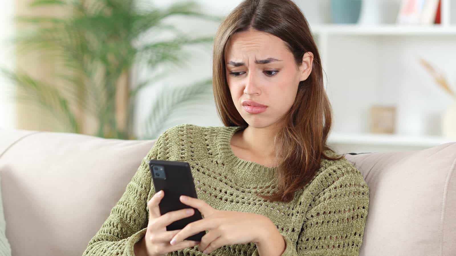 A woman sitting on a sofa looking at her smartphone with a concerned expression.