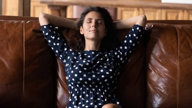 A woman in a navy blue polka-dot dress relaxing with her eyes closed and hands behind her head on a leather sofa.