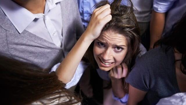 A woman with a distressed expression looks upward, clutching her hair while surrounded by a crowd of people.