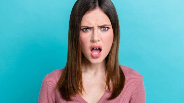 A young woman with a shocked expression, her mouth slightly open, against a turquoise background.