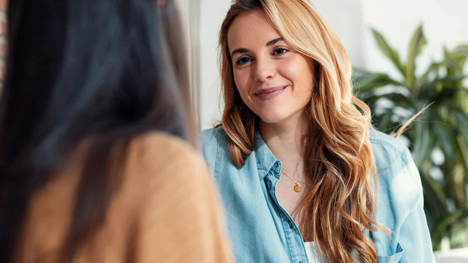 A woman with long wavy blonde hair smiles while looking at a person sitting across from her.