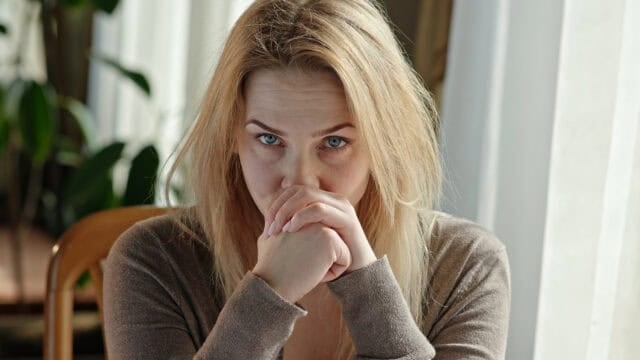 A woman looking forward with a serious expression, resting her chin on her clasped hands.