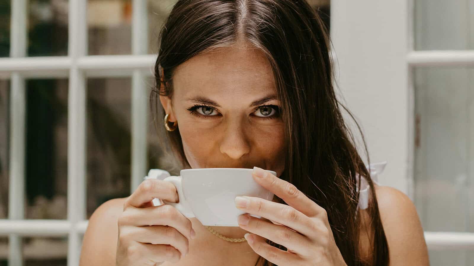 A woman holding a white coffee cup to her mouth and looking directly at the camera.