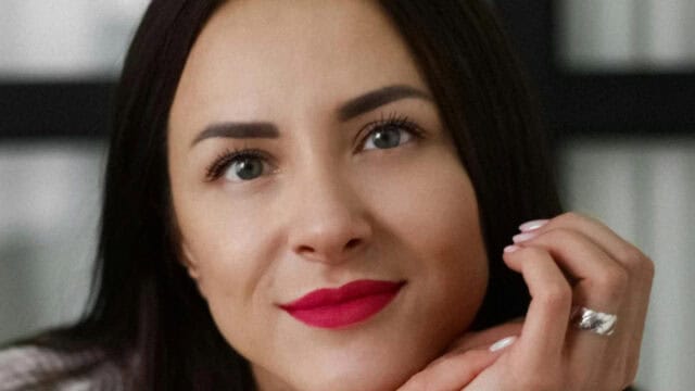 Close-up portrait of a woman with dark hair and bright red lipstick, resting her chin on her hand.