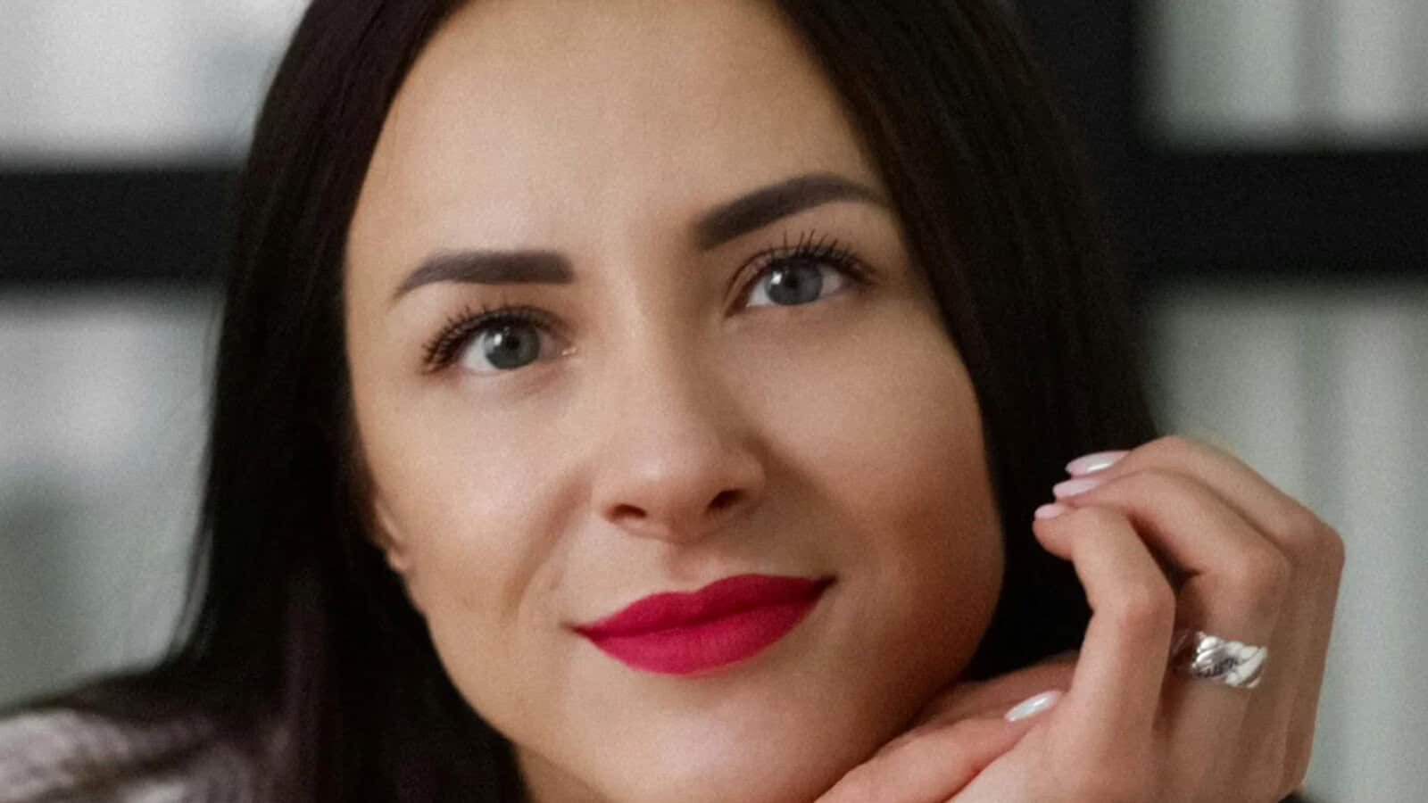 Close-up portrait of a woman with dark hair and bright red lipstick, resting her chin on her hand.