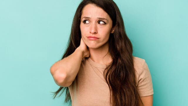 A young woman with long brown hair wearing a tan t-shirt looks to the side with a pensive expression, touching the back of her neck.
