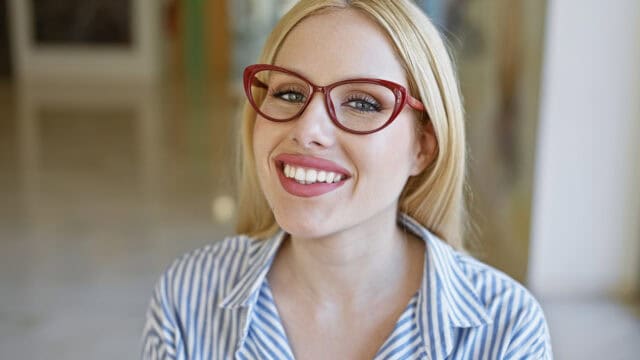 A smiling blonde woman wearing red cat-eye glasses and a blue and white striped shirt.