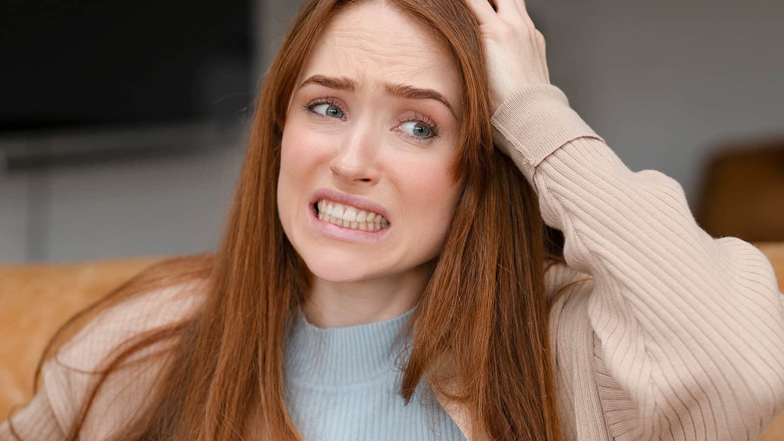 A woman with long red hair grimacing with her hand on her head.