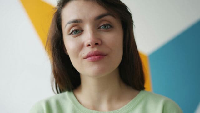 A woman with light eyes and long dark hair looks directly at the camera.