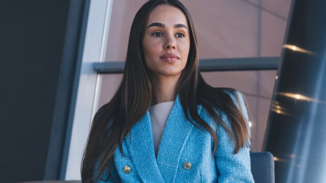 A woman with long dark hair wearing a bright blue double-breasted blazer sits near a window.
