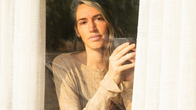 A woman holds a glass while looking out through a window between sheer curtains.