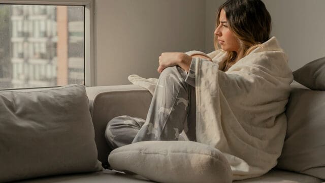 A woman in pajamas wrapped in a blanket sits on a couch, looking out a window.