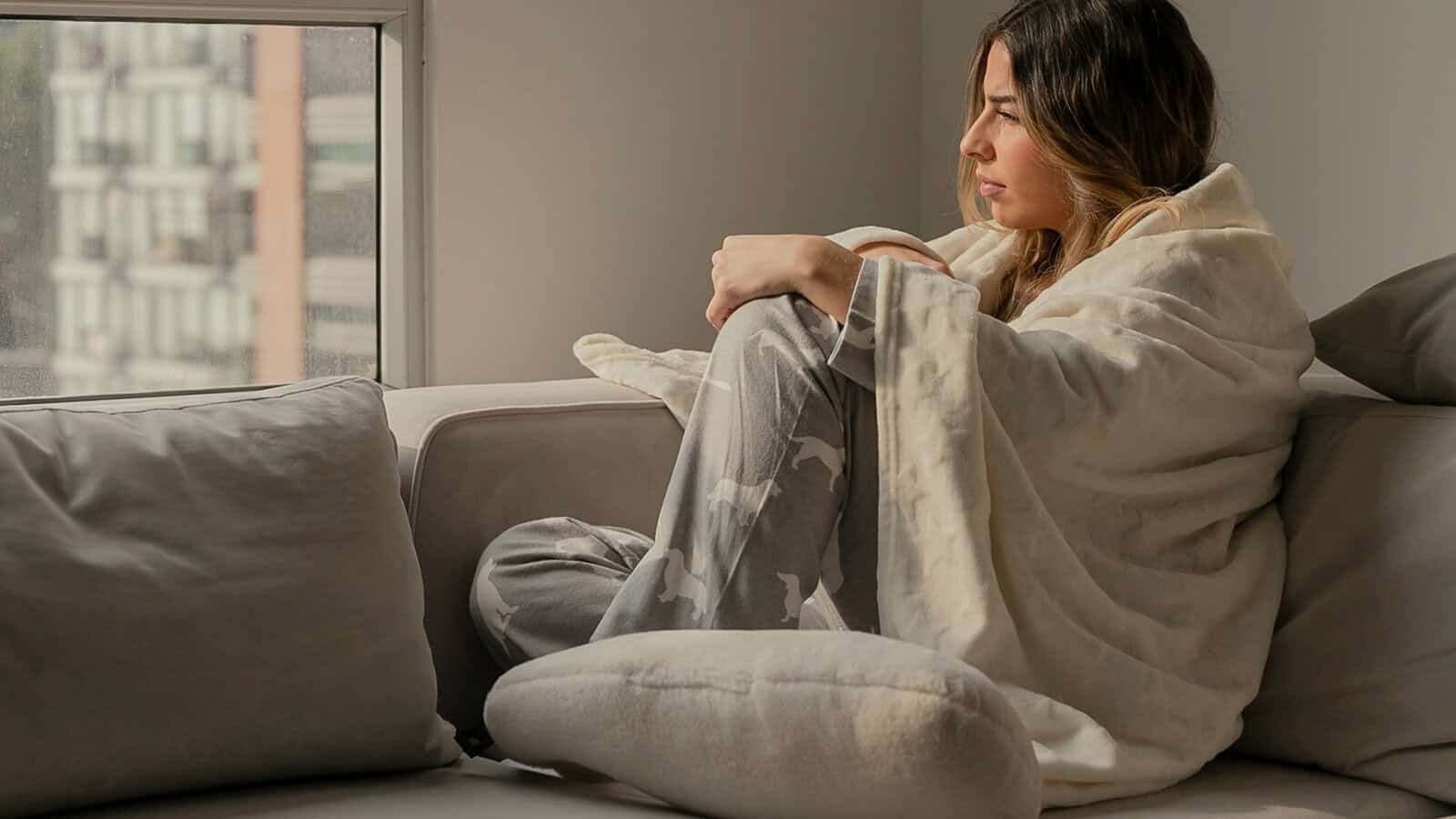 A woman in pajamas wrapped in a blanket sits on a couch, looking out a window.