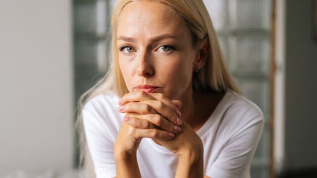 A woman with blonde hair looking directly at the camera with her hands clasped near her chin.