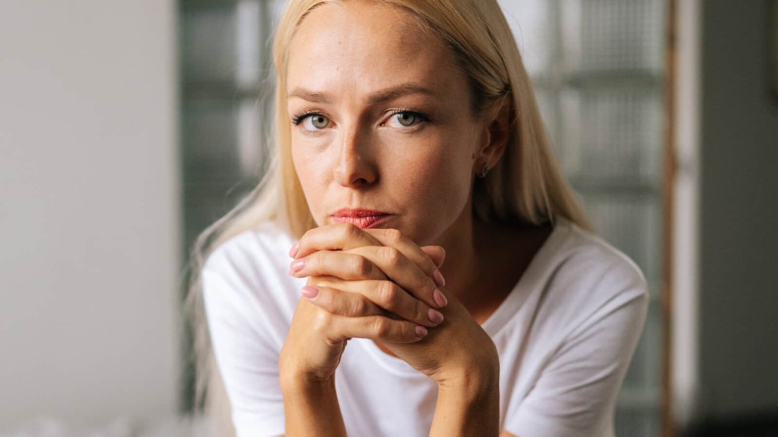 A woman with blonde hair looking directly at the camera with her hands clasped near her chin.