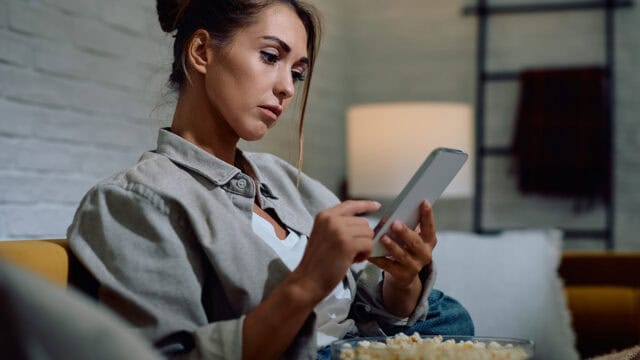 A woman sitting on a sofa looks at her smartphone while holding a bowl of popcorn.