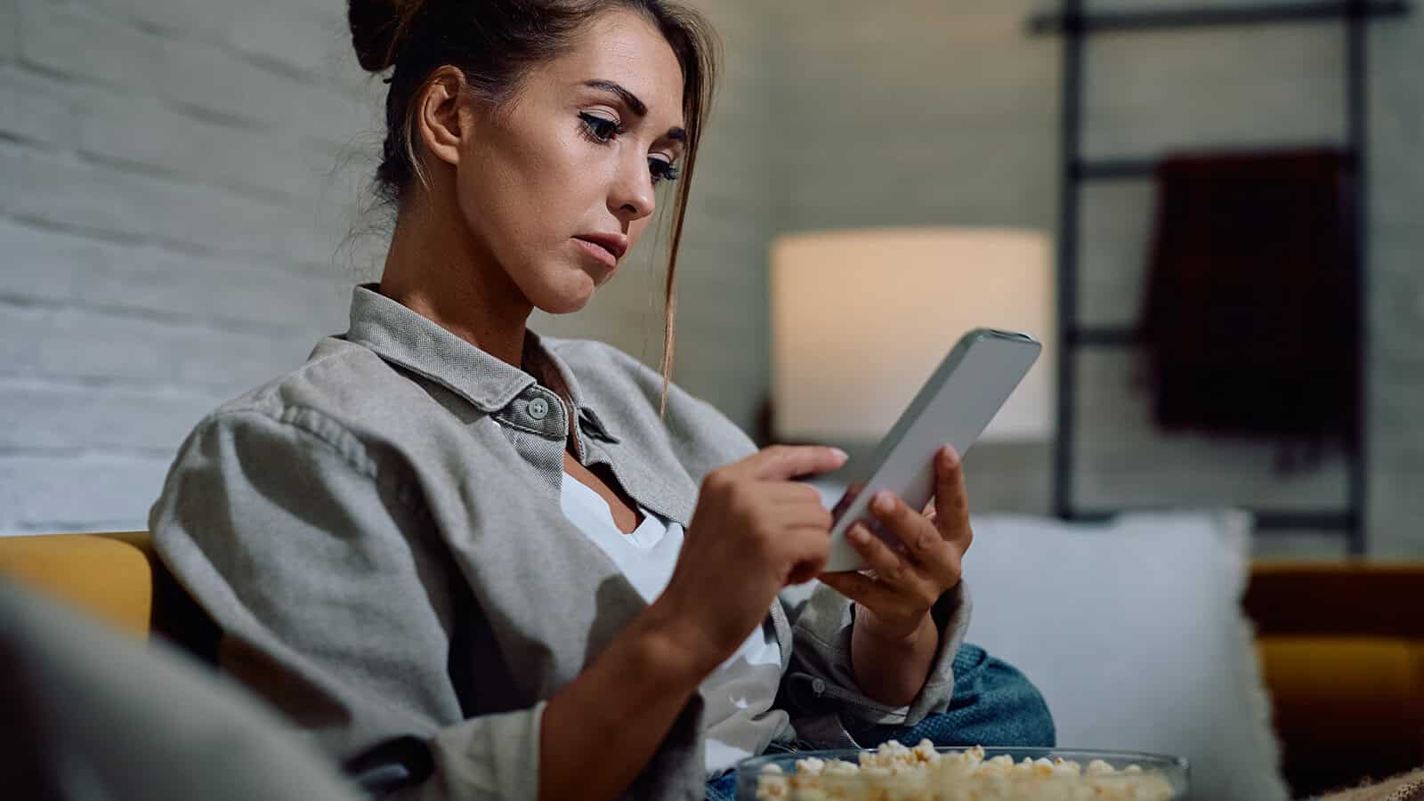 A woman sitting on a sofa looks at her smartphone while holding a bowl of popcorn.