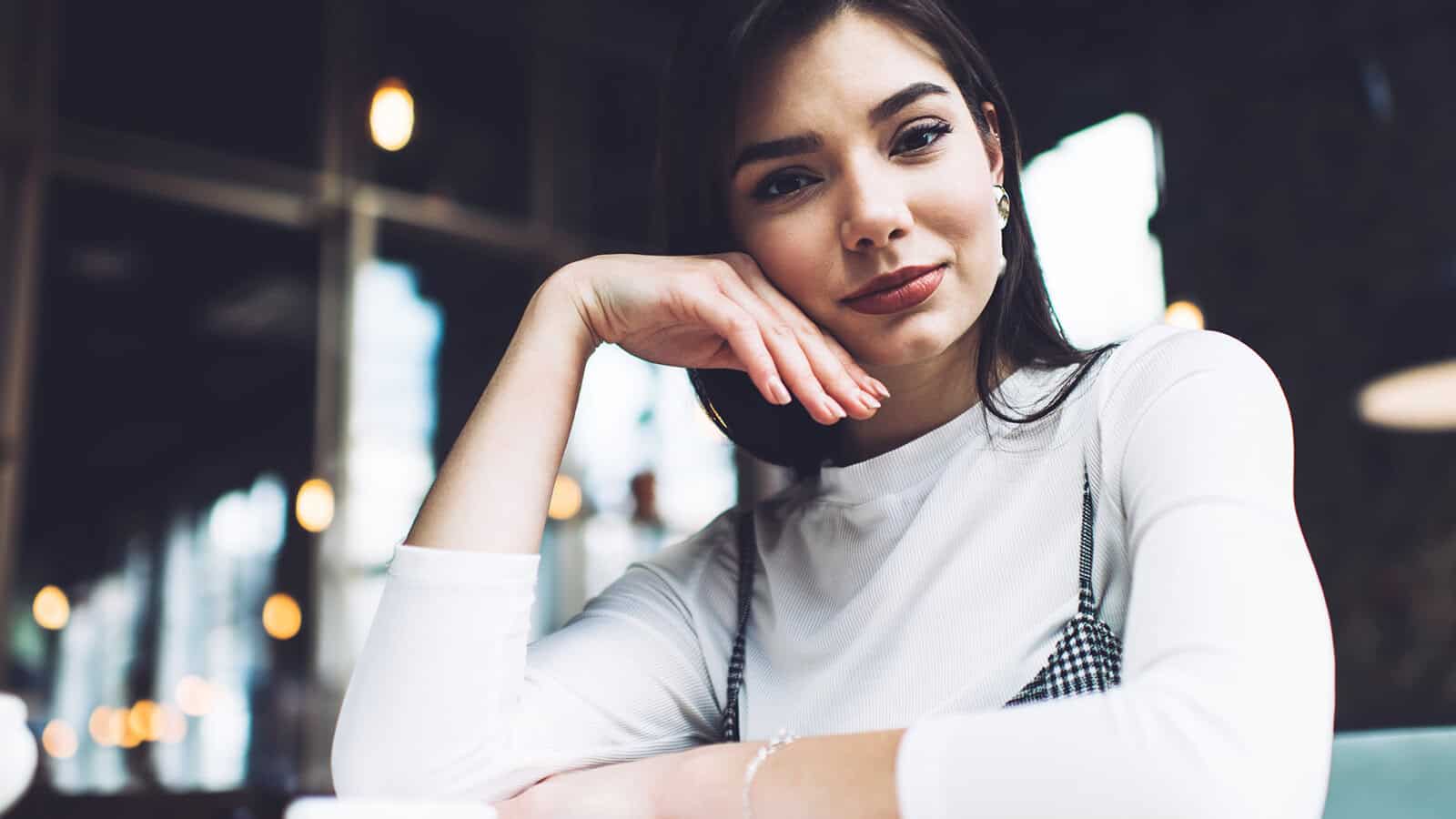 A woman with dark hair rests her chin on her hand, looking directly at the camera.