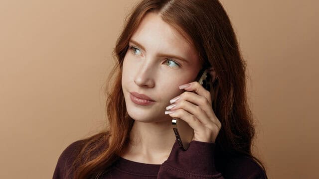 A woman with long red hair holds a smartphone to her ear.