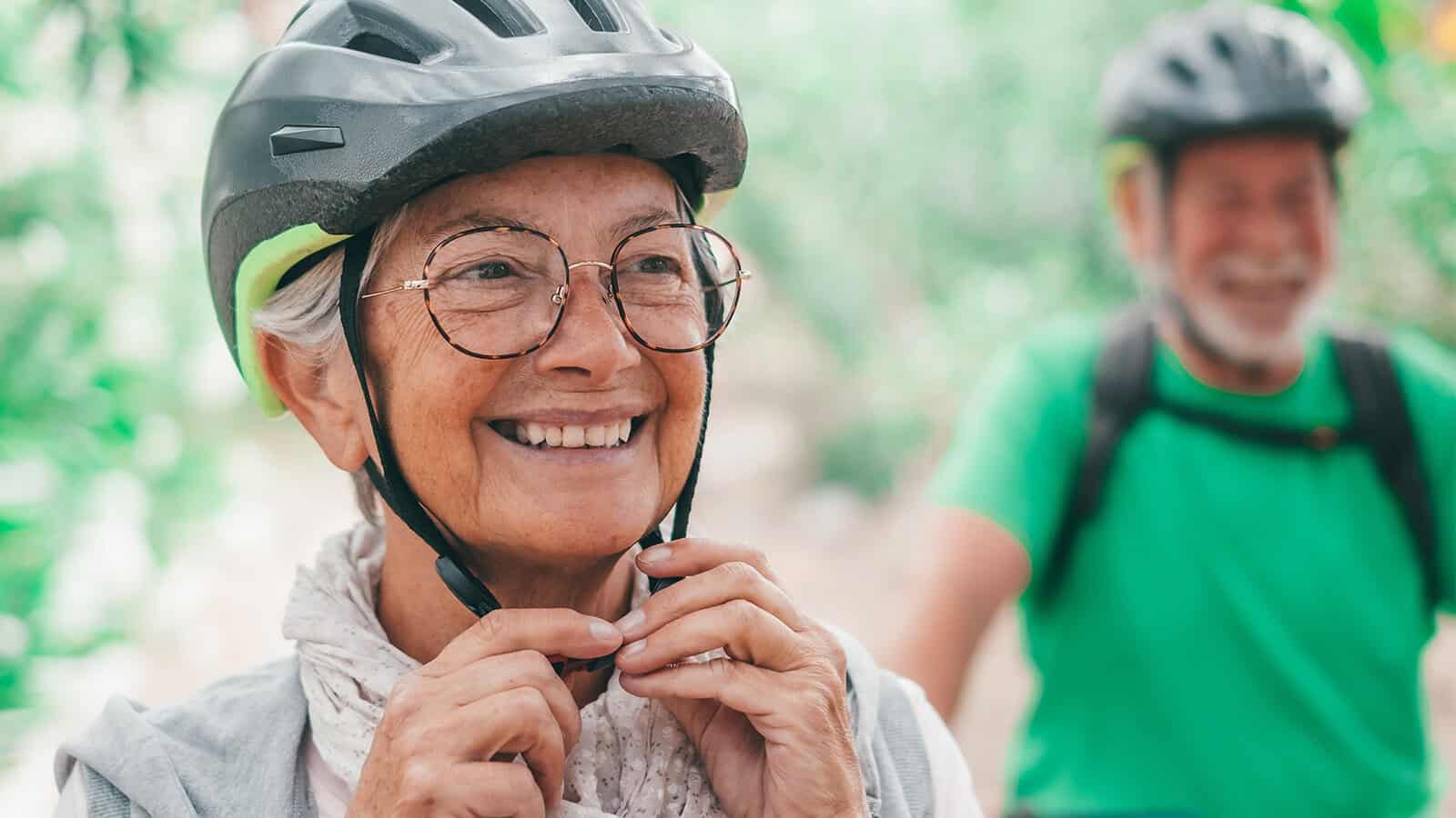 A smiling older woman in a cycling helmet adjusts her chin strap, with a man in a helmet and green shirt slightly out of focus in the background.