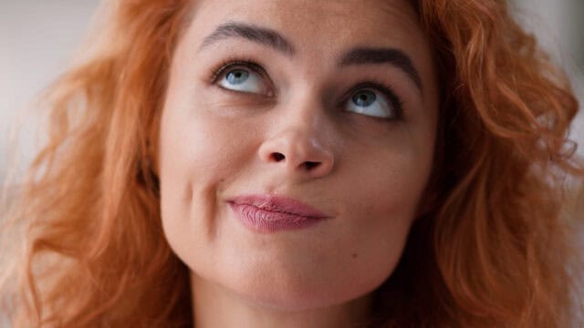 A person with curly red hair looking upward with a thoughtful, skeptical expression.