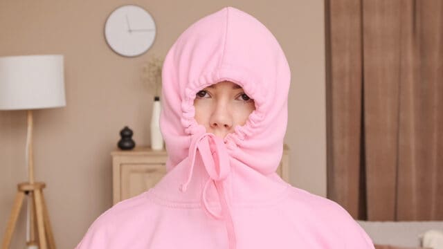 A person wearing a pink hooded sweatshirt with the drawstring pulled tight around their face.