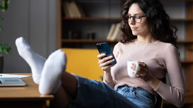A woman wearing glasses and a pink long-sleeved shirt sits at a desk with her feet in white socks resting on the surface, looking at her smartphone while holding a mug.