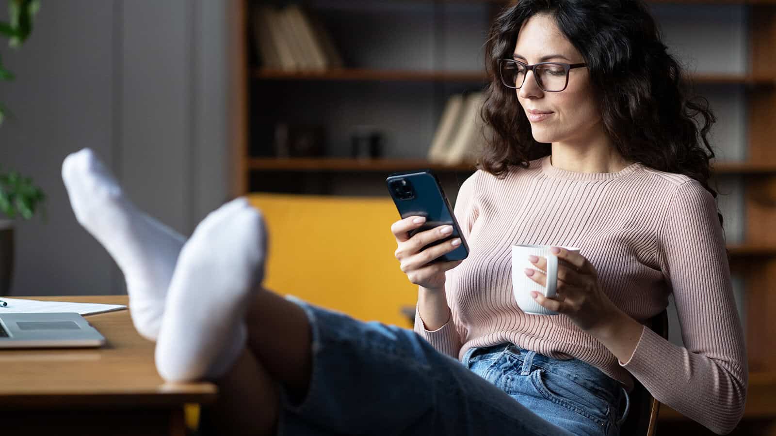 A woman wearing glasses and a pink long-sleeved shirt sits at a desk with her feet in white socks resting on the surface, looking at her smartphone while holding a mug.